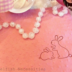 May include: A pink, textured gift bag with a bunny illustration. A clear beaded necklace is draped across the bag. The text "Selfish Necessities" is at the bottom of the image. Ribbons and a white plate are in the background.