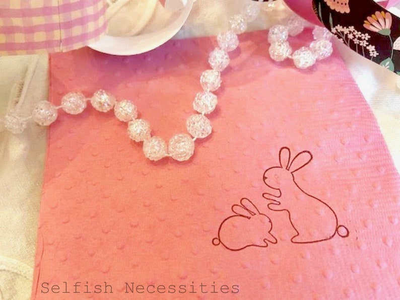 May include: A pink, textured gift bag with a bunny illustration. A clear beaded necklace is draped across the bag. The text "Selfish Necessities" is at the bottom of the image. Ribbons and a white plate are in the background.
