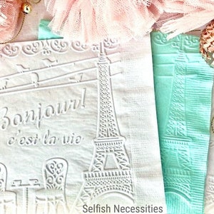 May include: Two white paper napkins with an embossed design of the Eiffel Tower and the text "Bonjour! c'est la vie". The napkins are on a pink and white background.