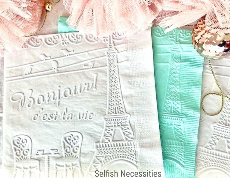 May include: Two white paper napkins with an embossed design of the Eiffel Tower and the text "Bonjour! c'est la vie". The napkins are on a pink and white background.