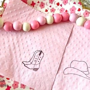 May include: Pink napkins with a dotted texture feature line drawings of a cowboy boot and hat. A beaded garland with pink and white wooden beads sits above the napkins. The words "Selfish Necessities" are in the bottom left corner.