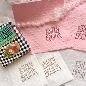 May include: Four white napkins with the text "and the story begins" printed in brown ink. Two pink napkins with the same text printed in black ink. A small black and white checkered book titled "Friendship, A Bouquet of Quotes".