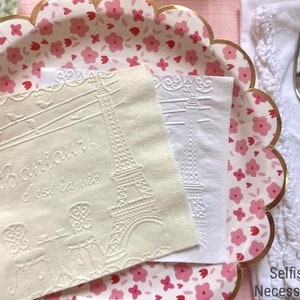 May include: Two white paper napkins with embossed designs. One napkin features the Eiffel Tower and the text "Bonjour! C'est la vie". The other napkin has a simple Eiffel Tower design. The napkins are on a pink and white floral paper plate with a gold scalloped edge.