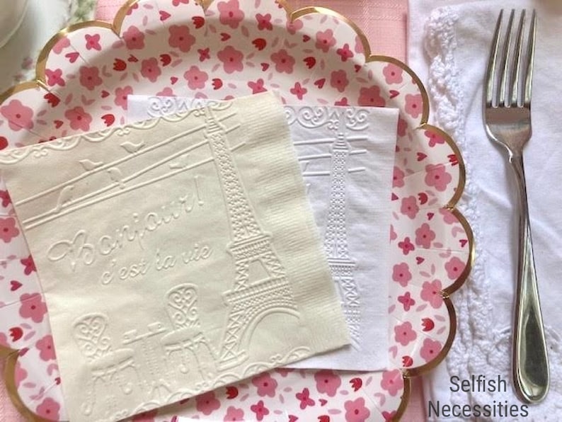 May include: Two white paper napkins with embossed designs. One napkin features the Eiffel Tower and the text "Bonjour! C'est la vie". The other napkin has a simple Eiffel Tower design. The napkins are on a pink and white floral paper plate with a gold scalloped edge.