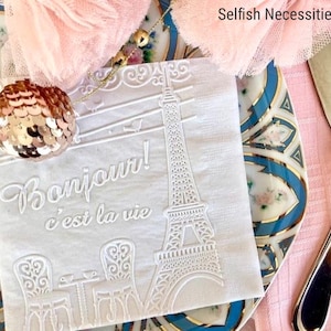 May include: White paper napkin with an embossed design of the Eiffel Tower, a table with two chairs, and the text "Bonjour! c'est la vie". The napkin is on a blue and white patterned plate.