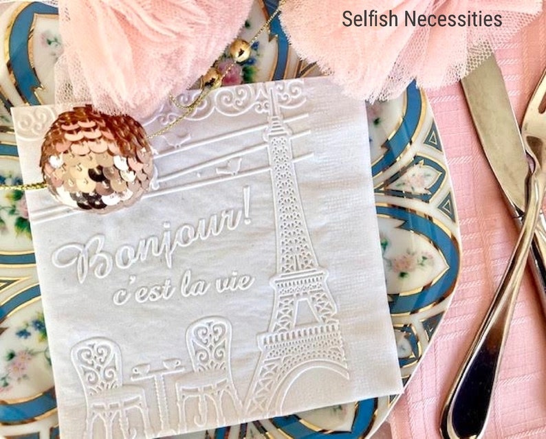 May include: White paper napkin with an embossed design of the Eiffel Tower, a table with two chairs, and the text "Bonjour! c'est la vie". The napkin is on a blue and white patterned plate.