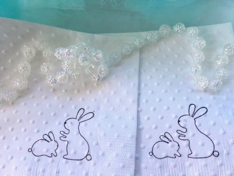 Mama and baby bunny image hand stamped on beverage size napkins in white with raised dots to provide texture. Mama bunny is sitting up with hands outreached to the baby bunny. The bunnies are facing each other.