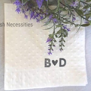 May include: A square white paper napkin with a subtle embossed heart pattern throughout. The text Selfish Necessities is printed in the upper left corner, and the letters B and D separated by a heart symbol are printed in the lower center.