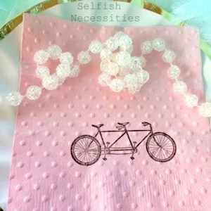 May include: A pink napkin with a dotted pattern and a tandem bicycle illustration. A white beaded garland is draped across the napkin, which rests on a gold-rimmed plate. The text "Selfish Necessities" is at the top.