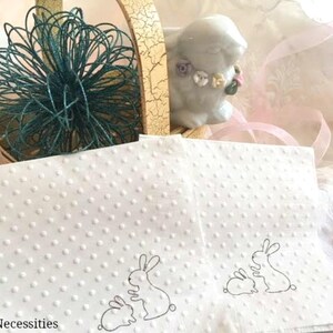 May include: White napkins with a dotted pattern and bunny illustrations are displayed with a ceramic bunny figurine and a decorative basket. A teal wire decoration sits atop the basket. The text "Selfish Necessities" is visible.