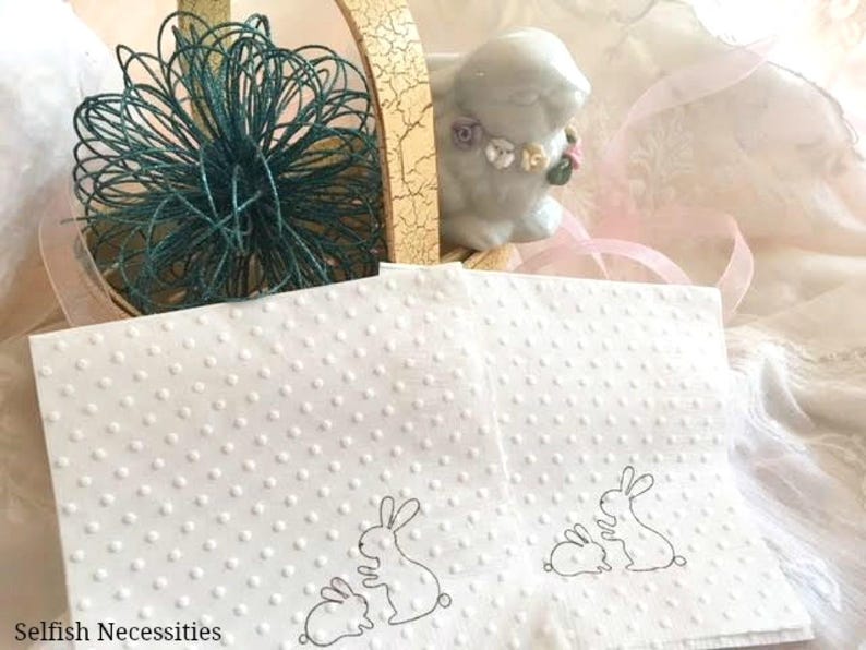 May include: White napkins with a dotted pattern and bunny illustrations are displayed with a ceramic bunny figurine and a decorative basket. A teal wire decoration sits atop the basket. The text "Selfish Necessities" is visible.