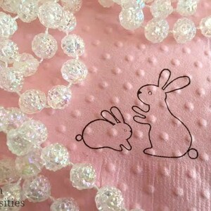 May include: A pink, textured surface with a beaded garland and two outlined bunny illustrations. The garland is made of clear, iridescent, spherical beads. The bunnies are black outlines, one sitting and one standing.