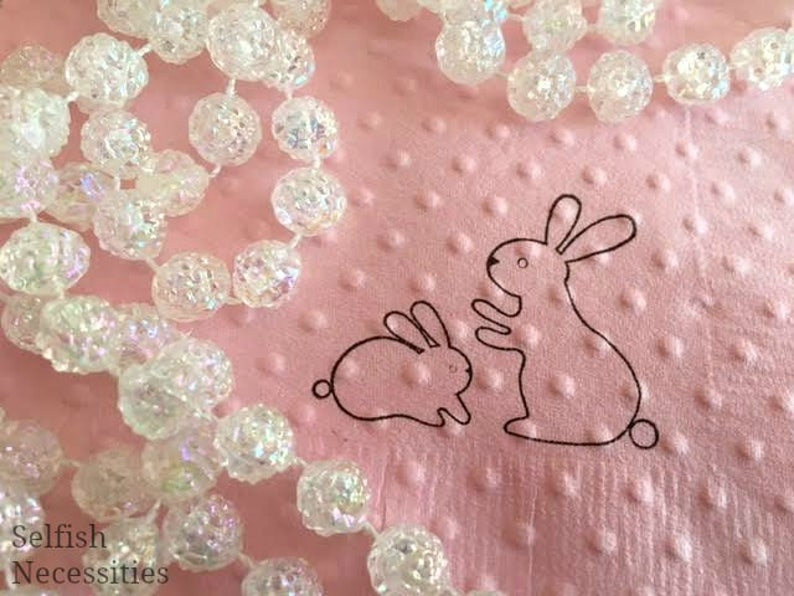 May include: A pink, textured surface with a beaded garland and two outlined bunny illustrations. The garland is made of clear, iridescent, spherical beads. The bunnies are black outlines, one sitting and one standing.