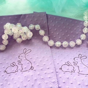 May include: Two lavender napkins with a dotted texture and a black outline of a mother and baby bunny. A strand of clear, beaded garland is draped across the napkins, with a light green and blue background.