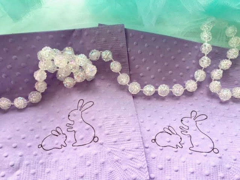 May include: Two lavender napkins with a dotted texture and a black outline of a mother and baby bunny. A strand of clear, beaded garland is draped across the napkins, with a light green and blue background.