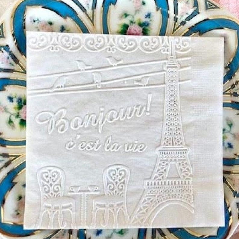 Cocktail size napkin embossed with a french scene featuring a cafe table and chair with coffee, the word Bonjour and the eiffel tower.