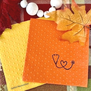 May include: Two square paper napkins in yellow and orange with a raised dot texture. The orange napkin features a black line drawing of a stethoscope shaped into a heart. The text Selfish Necessities is printed in black on the yellow napkin.