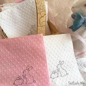 May include: A stack of pink and white dotted napkins with a bunny design. A ceramic bunny figurine with a blue bow sits nearby. The napkins are part of a set, perfect for Easter or spring celebrations.