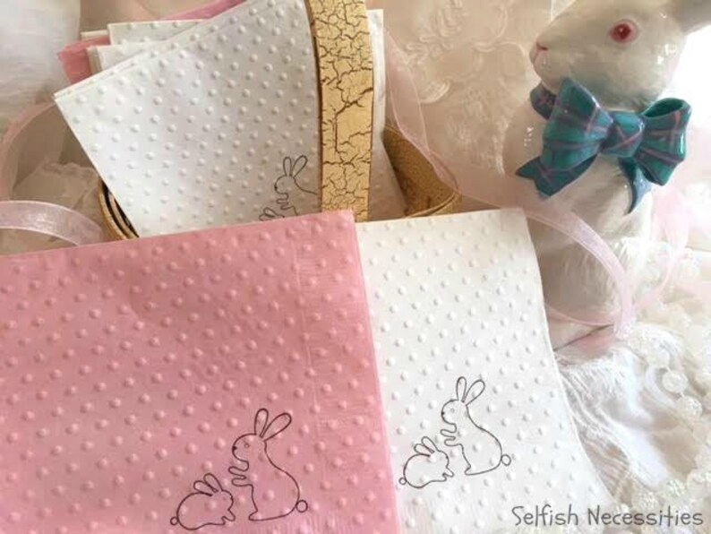 May include: A stack of pink and white dotted napkins with a bunny design. A ceramic bunny figurine with a blue bow sits nearby. The napkins are part of a set, perfect for Easter or spring celebrations.