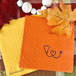 May include: Two square paper napkins, one yellow and one orange, featuring a raised dot texture. The orange napkin displays a black line drawing of a stethoscope shaped into a heart. The text Selfish Necessities is printed in black on the yellow napkin.