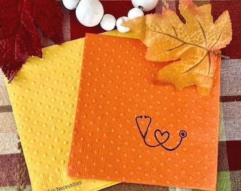 May include: Two square paper napkins, one yellow and one orange, featuring a raised dot texture. The orange napkin displays a black line drawing of a stethoscope shaped into a heart. The text Selfish Necessities is printed in black on the yellow napkin.