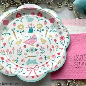 May include: A set of pastel pink and green paper plates with a floral and animal print. The plates feature a variety of flowers, bunnies, ducks, and mushrooms. The plates are decorated with a light blue border. The plates are part of a set of party supplies with the text "Our Story Begins" on a pink napkin.