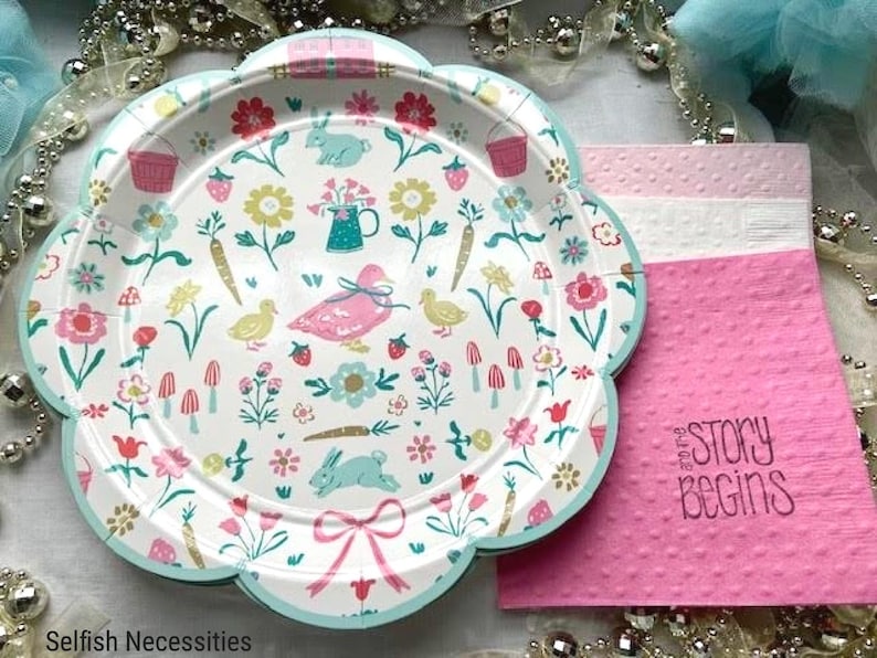 May include: A set of pastel pink and green paper plates with a floral and animal print. The plates feature a variety of flowers, bunnies, ducks, and mushrooms. The plates are decorated with a light blue border. The plates are part of a set of party supplies with the text "Our Story Begins" on a pink napkin.
