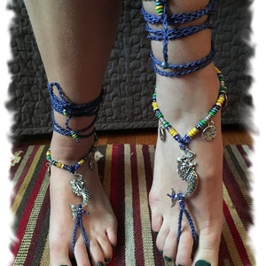 May include: Blue crocheted barefoot sandals with a mermaid charm and colorful beads. The sandals are worn on a pair of feet with dark green nail polish.