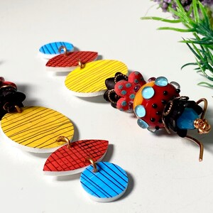 May include: A pair of colorful earrings with geometric shapes and a beaded charm. The earrings are made of white, yellow, red, and blue. The charm is made of red, yellow, blue, and black beads.