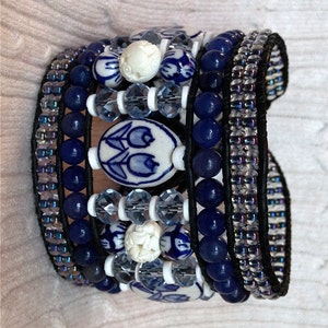 Puede incluir: Una pulsera envolvente de varias hebras con cuentas azules y blancas, cristales transparentes y cuentas blancas talladas. La pulsera tiene una base de cuero negro y está adornada con cuentas ovaladas de cerámica azul y blanca con un diseño de tulipanes.