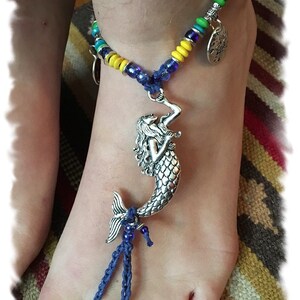 May include: A silver mermaid charm hangs from a blue and yellow beaded anklet. The anklet is tied around a person's ankle.