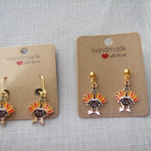 May include: Two pairs of gold-toned metal earrings featuring enamel turkey charms with orange, yellow, and brown feathers. Each pair is attached to a rectangular brown cardboard display card printed with the text Handmade with love.