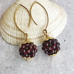 May include: A pair of gold-colored drop earrings. Each earring features a cluster of dark red beads, resembling grapes, suspended from a gold-colored hook. The earrings are displayed on a seashell.