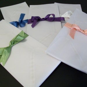 May include: Five white envelopes with different colored satin ribbons tied in bows. The ribbons are green, blue, purple, pink, and white.