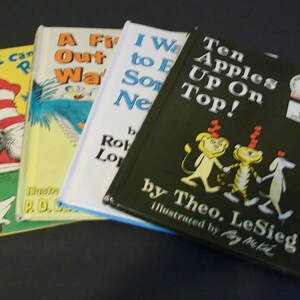 May include: Four children's books stacked on top of each other. The top book is green with the title "Ten Apples Up On Top!" by Theo. LeSieg, illustrated by Roy McKie. The second book is white with the title "I Want to Eat Something New!" by Robert Munsch. The third book is yellow with the title "A Fish Out of Water" by P.D. Eastman. The bottom book is yellow with the title "I Can Read With My Eyes Shut!" by Dr. Seuss.