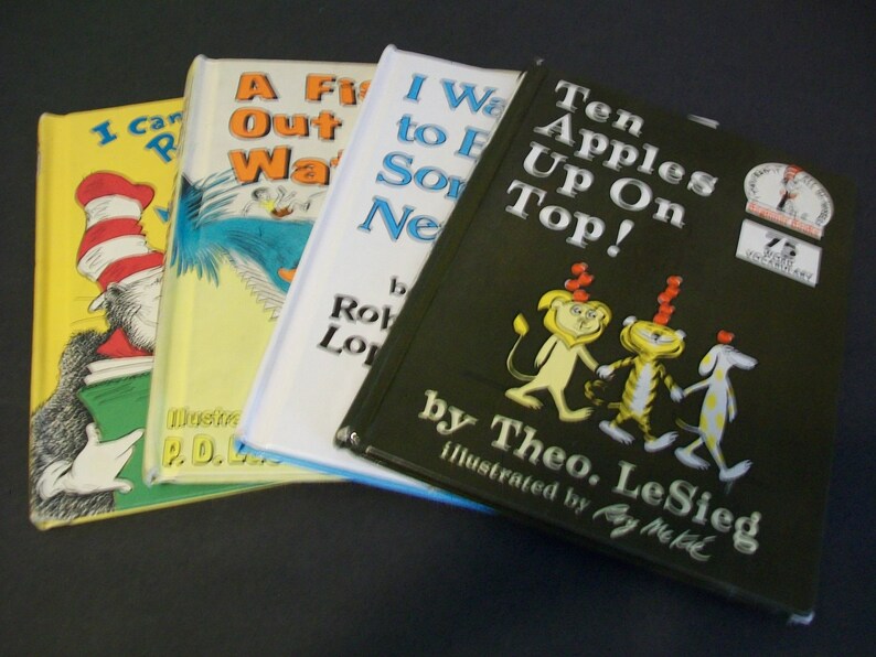 May include: Four children's books stacked on top of each other. The top book is green with the title "Ten Apples Up On Top!" by Theo. LeSieg, illustrated by Roy McKie. The second book is white with the title "I Want to Eat Something New!" by Robert Munsch. The third book is yellow with the title "A Fish Out of Water" by P.D. Eastman. The bottom book is yellow with the title "I Can Read With My Eyes Shut!" by Dr. Seuss.