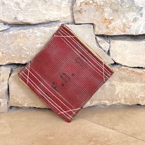 Puede incluir: Un libro rojo rectangular, cubierto de tela con costuras blancas y un borde amarillo. El libro est&aacute; sobre una superficie de piedra marr&oacute;n claro.