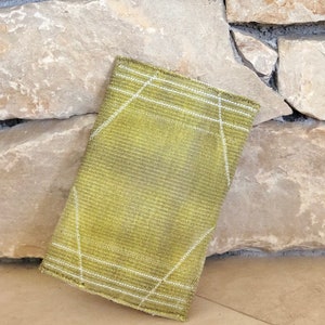 Puede incluir: Una bolsa de tejido verde tejido con l&iacute;neas blancas. La bolsa est&aacute; doblada y apoyada contra una pared de piedra.
