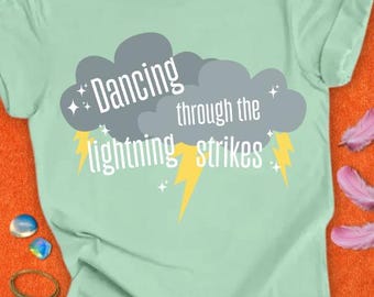 May include: A light green t-shirt with the text "Dancing through the lightning strikes" in white and yellow. The design features gray clouds and yellow lightning bolts. The shirt is made of a soft material.