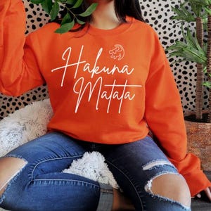 Peut inclure: Sweat-shirt orange avec l'inscription "Hakuna Matata" en écriture blanche et un petit symbole du roi lion au-dessus du texte.