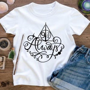 Puede incluir: Camiseta blanca con un gráfico negro de "Always", que incorpora el símbolo de la plataforma 9 3/4. La imagen incluye pantalones cortos vaqueros azules y una varita de madera. La camiseta está sobre una superficie de madera.