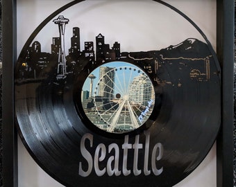 May include: A black vinyl record with a white silhouette of the Seattle skyline, including the Space Needle, and a photo of the Seattle Ferris wheel. The record is framed in a black frame and the word "Seattle" is printed in white on the record.