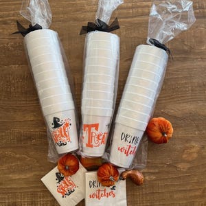 May include: Three sets of white Halloween-themed cups wrapped in cellophane with black bows. Each cup has a different orange and black graphic and text: "Cast a Spell", "Treat", and "Drink Witches". Small orange pumpkins and napkins are also included.