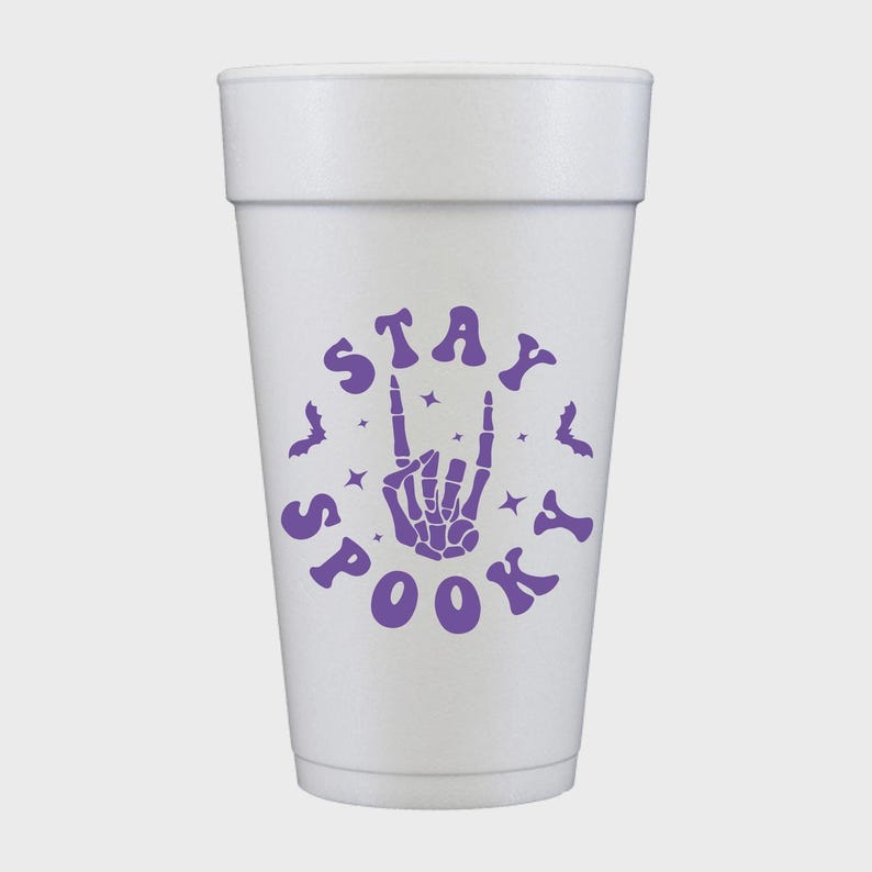 May include: A white foam cup with a purple "Stay Spooky" design. The design includes a skeleton hand making a rock-and-roll gesture, bats, and stars. The cup is cylindrical and has a slightly tapered shape.