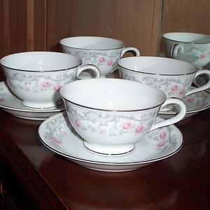 May include: Set of five white teacups and saucers with a floral pattern in shades of pink, blue, and green. The cups and saucers have a silver trim.