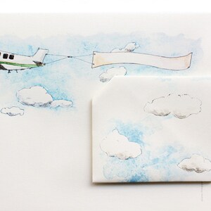 Pode incluir: Ilustração em aquarela de um pequeno avião puxando uma faixa em branco através de um céu azul com nuvens brancas e fofas. Um envelope correspondente com ilustrações de nuvens também é mostrado.
