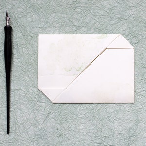 Pode incluir: Um envelope branco com uma aba dobrada, sobre uma superf&iacute;cie verde texturizada. Uma caneta de caligrafia preta est&aacute; &agrave; esquerda do envelope.