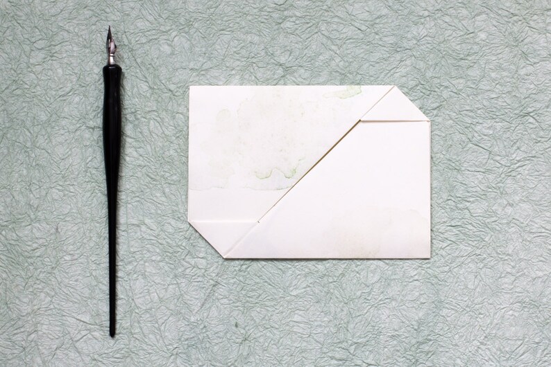 Pode incluir: Um envelope branco com uma aba dobrada, sobre uma superf&iacute;cie verde texturizada. Uma caneta de caligrafia preta est&aacute; &agrave; esquerda do envelope.