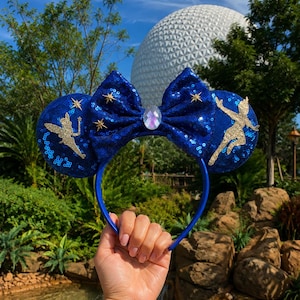 May include: A dark blue sequined headband featuring two round ears and a central bow. The left ear displays a gold glitter silhouette of a fairy, while the right ear shows a gold glitter silhouette of a flying boy. Small gold stars and a central iridescent gem accent the bow.