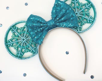 May include: A teal sequined bow and matching snowflake ears on a headband. The ears are round with a snowflake design and are embellished with rhinestones. The headband is light blue and brown. The background is white with scattered blue sequins.